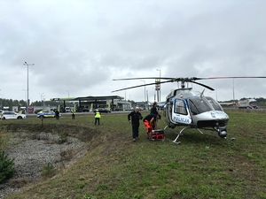 Śmigłowiec stoi na trawiastym terenie. Obok policjant i ratownik medyczny. W głębi stacja benzynowa i pojazdy na parkingu przy stacji benzynowej oraz radiowóz policyjny i policjanci.