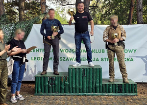 Na zdjęciu uczestnicy zawodów strzeleckich stoi na podium po zakończonym turnieju. Mężczyźni trzymają w ręku puchar.