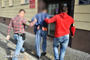 Dwaj nieumundurowani policjanci, w opaskach na ramieniu z napisem Policja, prowadzą zatrzymanego.