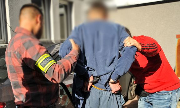 Dwaj nieumundurowani policjanci, w opaskach na ramieniu z napisem Policja, prowadzą zatrzymanego do radiowozu.