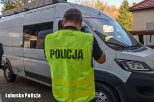 Na zdjęciu policjant w odblaskowej kamizelce z napisem Policja fotografuje odzyskanego białego busa, który stoi na posesji.