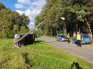 Miejsce zdarzenia. W rowie leży przewrócony samochód, obok zaparkowany radiowóz policyjny i samochód osobowy, przy którym stoją policjanci i mężczyźni.