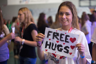 Uśmiechnięta dziewczyna prezentuje kartkę z wymalowanymi czerwonymi serduszkami i napisem w języku angielskim Free Hugs.