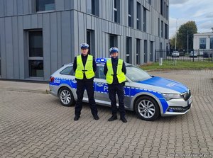 Dwaj policjanci w kamizelkach odblaskowych stojący koło radiowozu przed budynkiem policji.