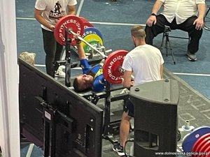 Na zdjęciu mężczyzna w trakcie wyciskania sztangi podczas zawodów. Obok ławeczki stoi dwóch mężczyzn, którzy asekurują sportowca.