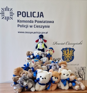 Sznupek i inne maskotki ułożone pod ścianką z napisem: Policja, Komenda Powiatowa Policji w Cieszynie, www.cieszyn.policja.gov.pl. Po prawej stronie widać herb Cieszyna i napis: Powiat Cieszyński.