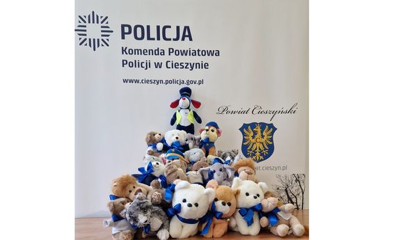 Sznupek i inne maskotki ułożone pod ścianką z napisem: Policja, Komenda Powiatowa Policji w Cieszynie, www.cieszyn.policja.gov.pl. Po prawej stronie widać herb Cieszyna i napis: Powiat Cieszyński.