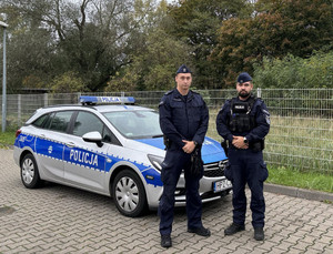 Na zdjęciu dwóch umundurowanych policjantów stoi przy radiowozie.