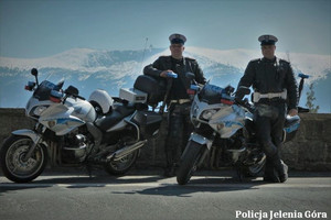 Dwaj umundurowani policjanci drogówki stoją przy motocyklach. W tle widać góry.