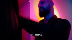 Stopklatka z filmu: Mężczyzna z brodą na tle ściany oświetlonej na żółto i różowo.