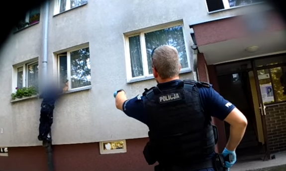 Policjant przed budynkiem mieszkalnym, drugi z policjantów wchodzi do mieszkania przez okno.