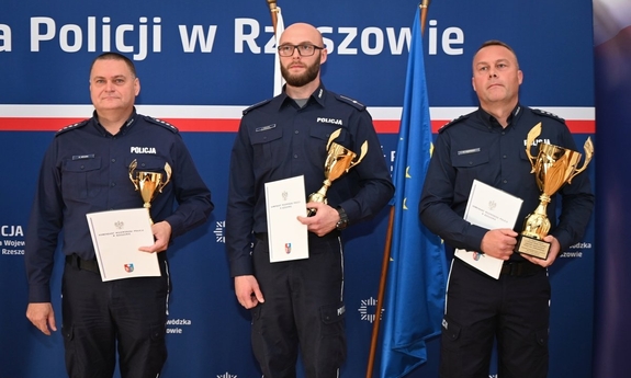 Trzej policjanci podczas konkursu służby dyżurnej w auli Komendy Wojewódzkiej Policji. Trzymają dyplomy i puchary.