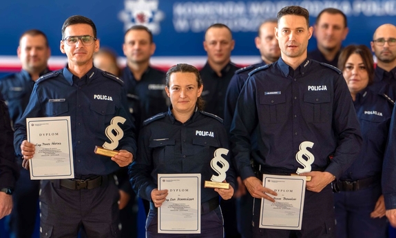 Grupa policjantów pozująca do zdjęcia grupowego. Policjantka i dwaj policjanci stojący obok niej trzymają dyplomy.