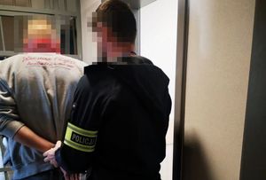 Na zdjęciu policjant z odblaskową naszywką na przedramieniu prowadzi zatrzymanego mężczyznę.