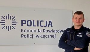 Na zdjęciu umundurowany policjant pozuje do zdjęcia. W tle napis na ścianie Policja, Komenda Powiatowa Policji w Łęcznej.