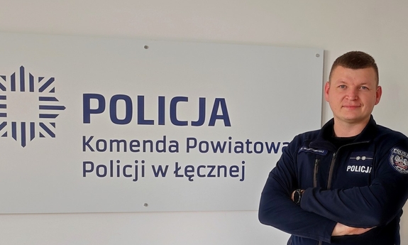 Na zdjęciu umundurowany policjant pozuje do zdjęcia. W tle napis na ścianie Policja, Komenda Powiatowa Policji w Łęcznej.