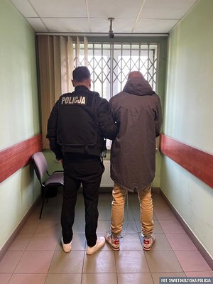 Na zdjęciu umundurowany policjant stoi z zatrzymanym mężczyzną na korytarzu budynku.