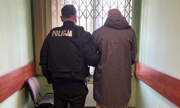 Na zdjęciu umundurowany policjant stoi z zatrzymanym mężczyzną na korytarzu budynku.