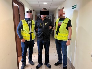 Dwaj policjanci z zatrzymaną osobą zakutą w kajdanki na korytarzu.