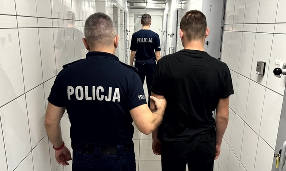 Dwóch umundurowanych policjantów prowadzących korytarzem zatrzymanego.