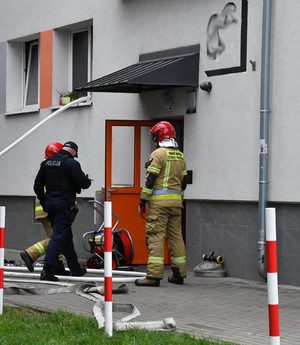 Policjant i dwaj strażacy przed budynkiem gdzie w jednym z mieszkań wybuchł pożar.