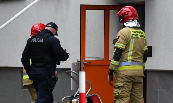 Policjant i dwaj strażacy przed budynkiem gdzie w jednym z mieszkań wybuchł pożar.