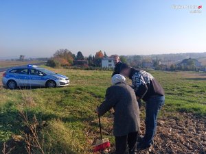 Na zdjęciu seniorka podpierająca się miotłą w objęciach syna. Policjant stoi z boku.