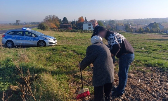 Na zdjęciu seniorka podpierająca się miotłą w objęciach syna. Policjant stoi z boku.