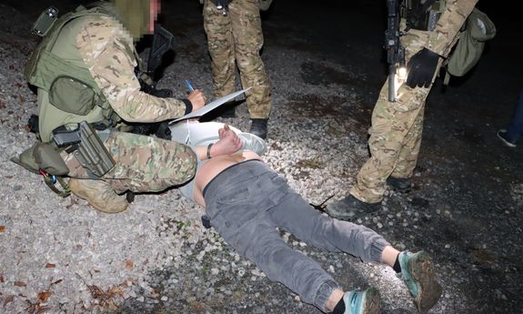 Pora nocna. Policjant kuca nad leżącym na ziemi zatrzymanym. Obok widoczne nogi dwóch innych policjantów.