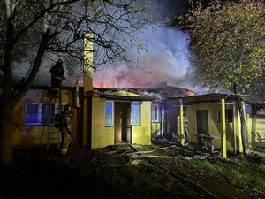 Na zdjęciu widok spalonego budynku mieszkalnego, obok dwóch strażaków w trakcie akcji gaśniczej.