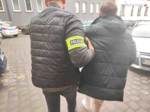 Policjant prowadzi zatrzymana osobę zakutą w kajdanki.