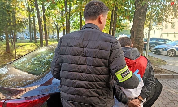 Policjant prowadzi zatrzymanego mężczyznę zakutego w kajdanki.