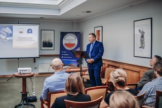 Mężczyzna wita uczestników spotkania, przed nim pulpit na notatki, na ekranie obok wyświetlana jest prezentacja.