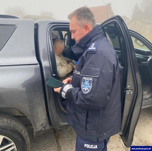 Na zdjęciu umundurowany policjant stoi przy otwartym samochodzie, w środku na tylnym siedzeniu pojazdu siedzi odnaleziona seniorka.