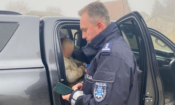 Na zdjęciu umundurowany policjant stoi przy otwartym samochodzie, w środku na tylnym siedzeniu pojazdu siedzi odnaleziona seniorka.