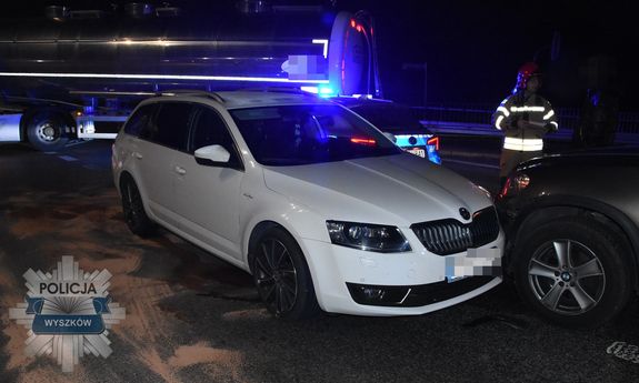 Pora nocna. Na pierwszym planie widoczna biała skoda, z prawej strony fragment auta, w które uderzyła skoda, z tyłu widać fragment policyjnego radiowozu i cysternę. Przy samochodach stoi strażak i osoba w ciemnym ubraniu.