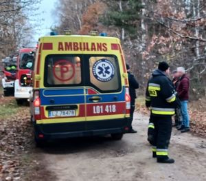 Karetka pogotowia na leśnej drodze, w tle wozy strażackie obok stoją mężczyźni.