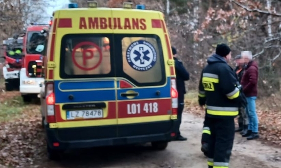 Karetka pogotowia na leśnej drodze, w tle wozy strażackie obok stoją mężczyźni.