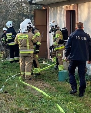 Fragment budynku przy którym stoją strażacy i umundurowany policjant.