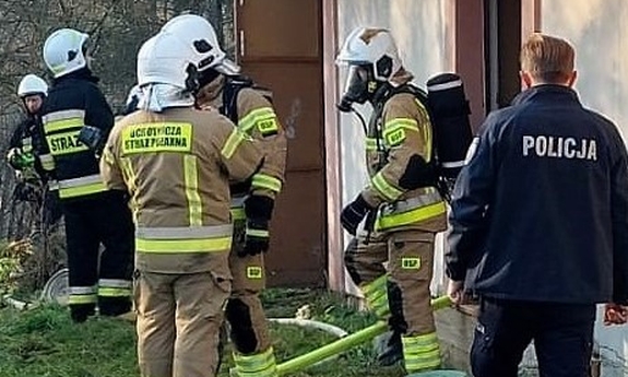 Fragment budynku przy którym stoją strażacy i umundurowany policjant.