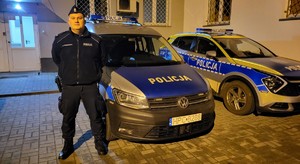 Policjant sierżant Grzegorz Dobaczewski stoi przed budynkiem obok dwóch zaparkowanych radiowozów.