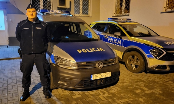 Policjant sierżant Grzegorz Dobaczewski stoi przed budynkiem obok dwóch zaparkowanych radiowozów.