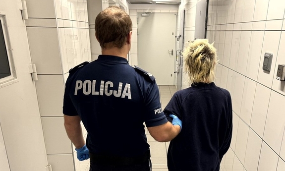 Umundurowany policjant prowadzący korytarzem zatrzymaną kobietę.