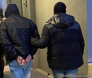 Policjant w ubraniu cywilnym prowadzi zatrzymanego skutego w kajdanki.