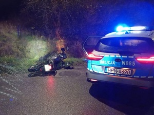 Motocykl leżący przy jezdni obok zaparkowany radiowóz z włączonymi sygnałami.