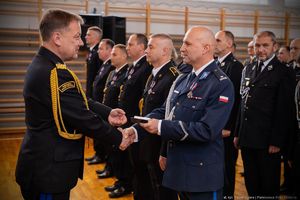 Na zdjęciu umundurowany policjant odbiera gratulacje od funkcjonariusza Państwowej Straży Pożarnej. W tle stoją funkcjonariusze Państwowej Straży Pożarnej biorący udział w uroczystości.