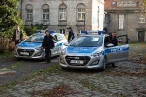 Trójka policjantów wysiadających z dwóch radiowozów na tle starego dworku.