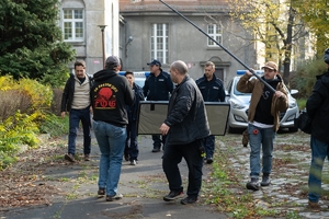 Plan filmowy, na pierwszym planie ekipa filmowa z kamerą oraz mikrofonem dalej policjanci i radiowozy na tle starego dworku.