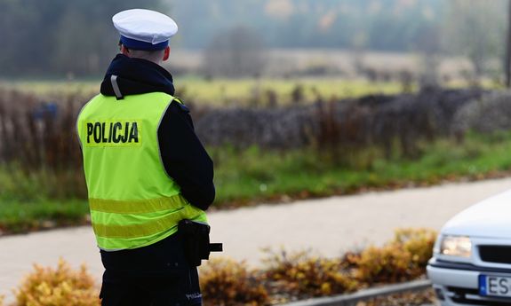 Na zdjęciu umundurowany policjant w żółtej kamizelce odblaskowej stoi naprzeciwko srebrnego pojazdu.