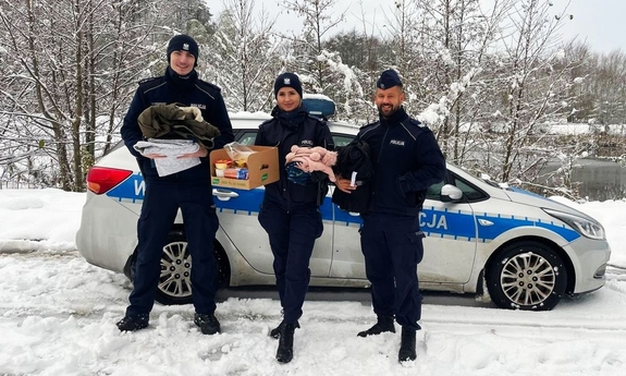 Trzej policjanci stoją przed radiowozem policyjnym, trzymają dary dal potrzebujących.